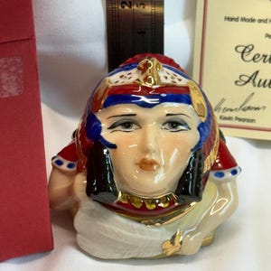 May include: A ceramic figurine of a person with a stylized headpiece in red, blue, and gold, featuring black hair accents and a white draped garment. A metal ruler is positioned behind the figurine, showing a height of approximately 2.5 inches.