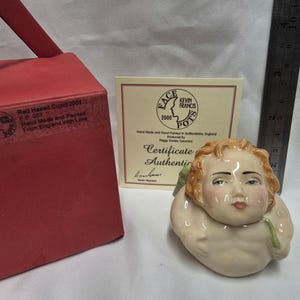 May include: A ceramic Face Pot figurine of a cupid with red hair, accompanied by a red box labeled Red Haired Cupid 2001, a certificate of authenticity, and a metal ruler showing a height of approximately 3.5 inches for the figurine.
