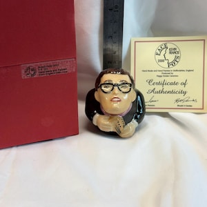 May include: A ceramic character jug of Buddy Holly wearing black-rimmed glasses, a black jacket, and a pink shirt. A metal ruler shows the figure is approximately 3.5 inches tall. Includes a red box and a Certificate of Authenticity from Face Pots.