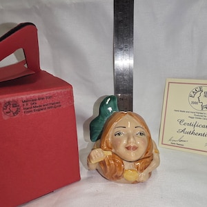 May include: A ceramic mermaid figurine with red hair and a green tail fin. The figurine is next to a red box and a certificate of authenticity. The box has text that reads "Mermaid Ariel 2001".