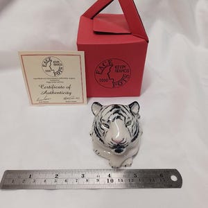 May include: A ceramic tiger figurine with black stripes and a pink nose, accompanied by a red box with a handle and a certificate of authenticity. The figurine is approximately 4 inches long.