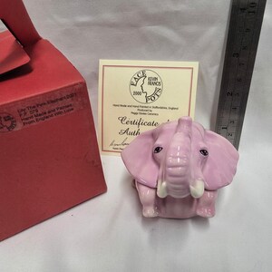 May include: A pink ceramic elephant figurine with white tusks, accompanied by a red box labeled Lily The Pink Elephant 2001, a certificate of authenticity, and a metal ruler showing a height of approximately 3 inches for the figurine.