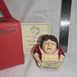 May include: A ceramic face pot figurine featuring a person with brown hair wearing a maroon garment, holding a small house model. It is accompanied by a certificate of authenticity, a red box, and a metal ruler showing a height of 3 inches.
