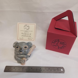 Puede incluir: Una figura de koala de cerámica sosteniendo una rama, con una combinación de colores gris y beige. Una caja roja con el texto "ACE KEVIN FRANCIS POTS 2000" está a la derecha. Un certificado de autenticidad está a la izquierda. Una regla metálica muestra 15 cm.