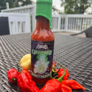 Pode incluir: Uma garrafa de molho picante com uma tampa verde e um rótulo preto que diz "Greenbrier Extreme" e "Ghost Pepper". A garrafa está cercada por pimentas fantasmas vermelhas.