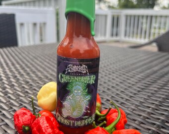 Może przedstawiać: Butelka ostrego sosu z zieloną zakrętką i czarną etykietą z napisem "Greenbrier Extreme" i "Ghost Pepper". Butelka jest otoczona czerwonymi papryczkami ghost pepper.