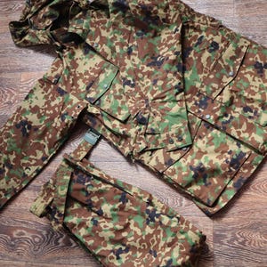 Puede incluir: Un conjunto de chaqueta y pantalón de camuflaje. La chaqueta tiene capucha y cierre de cremallera. El pantalón tiene trabillas para el cinturón y cierre de cremallera.