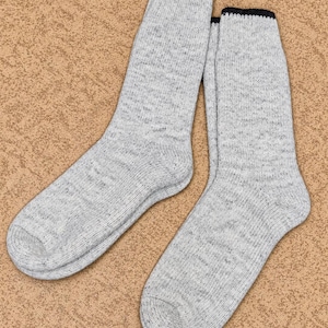 May include: A pair of light gray heathered knit socks featuring a subtle speckled texture throughout the fabric. Each sock is finished with a distinct, solid black ribbed cuff at the top, providing a clear contrast to the light gray body.