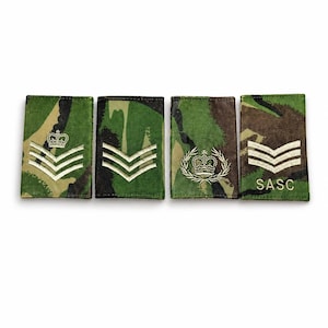 Puede incluir: Cuatro parches militares con fondos de camuflaje. Cada parche presenta galones blancos, coronas o el texto "SASC". Los parches son rectangulares y tienen una combinación de colores verde, marrón y negro.