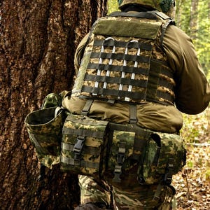 Könnte beinhalten: Eine Person trägt einen Tarnhelm und eine taktische Weste mit mehreren Taschen und Gurten. Die Weste und die Taschen sind in einem grün-braunen Tarnmuster gehalten, geeignet für Outdoor-Aktivitäten.