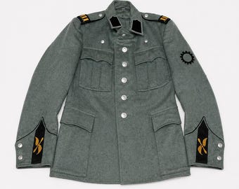 Puede incluir: Chaqueta militar gris con botones plateados, cuatro bolsillos delanteros y detalles en negro y dorado. La chaqueta presenta parches negros en el cuello, charreteras y un emblema de engranaje en la manga izquierda. Los puños tienen diseños en negro y dorado.