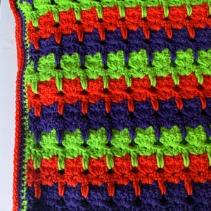 May include: A handmade crochet blanket with a vibrant color scheme. The blanket features rows of bright green, orange, and purple, with an orange border. The crochet pattern creates a textured, visually appealing design.