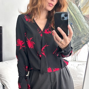 Peut inclure: Un chemisier noir à manches longues avec un imprimé floral rouge et un col en V. Le chemisier a une coupe enveloppante et une ceinture à la taille.