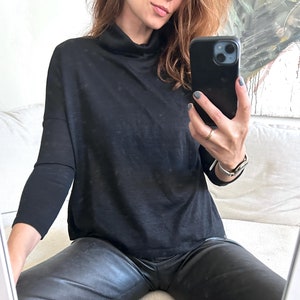 Puede incluir: Una mujer con una camisa negra de cuello alto y pantalones de cuero negro. Ella sostiene un teléfono inteligente negro en su mano derecha.