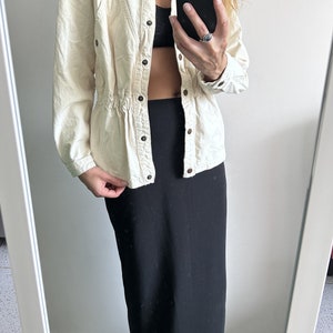 K&ouml;nnte beinhalten: Eine wei&szlig;e Jeansjacke mit eingezogenem Bund und einem schwarzen Maxirock. Die Jacke hat einen Knopfschlie&szlig;er und einen Stehkragen.