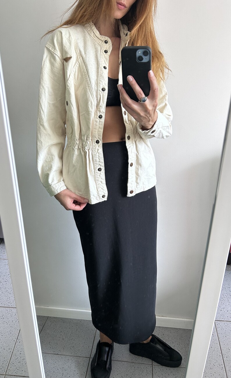 K&ouml;nnte beinhalten: Eine wei&szlig;e Jeansjacke mit eingezogenem Bund und einem schwarzen Maxirock. Die Jacke hat einen Knopfschlie&szlig;er und einen Stehkragen.
