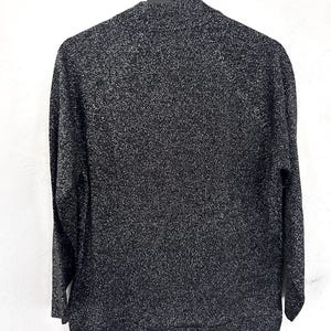 May include: A black and silver speckled long-sleeved sweater hangs on a black hanger. The sweater has a high neckline and a textured appearance. The fabric appears to be knit or woven, with a subtle sheen. The garment is displayed against a white background.