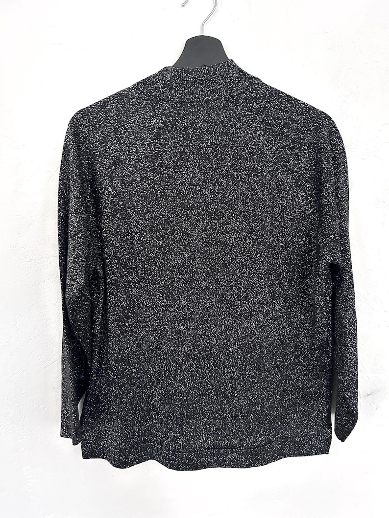May include: A black and silver speckled long-sleeved sweater hangs on a black hanger. The sweater has a high neckline and a textured appearance. The fabric appears to be knit or woven, with a subtle sheen. The garment is displayed against a white background.