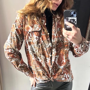 May include: A button-down shirt with a paisley pattern in shades of brown, orange, and white. The shirt has two chest pockets and is tied at the waist. The long-sleeved shirt is made of a soft, possibly velvet-like material.
