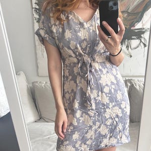 May include: A light blue floral print dress with a V-neckline and short sleeves. The dress features a tie waist and a knee-length hemline. The floral pattern is a cream color, and the dress is made of a lightweight fabric.
