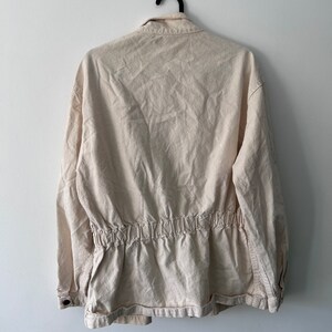 K&ouml;nnte beinhalten: Eine cremefarbene, lang&auml;rmelige Jacke mit einem Kordelzug in der Taille. Die Jacke hat einen Knopfleistenverschluss und einen Kragen.