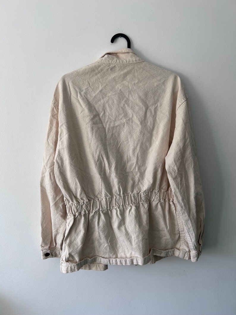 K&ouml;nnte beinhalten: Eine cremefarbene, lang&auml;rmelige Jacke mit einem Kordelzug in der Taille. Die Jacke hat einen Knopfleistenverschluss und einen Kragen.