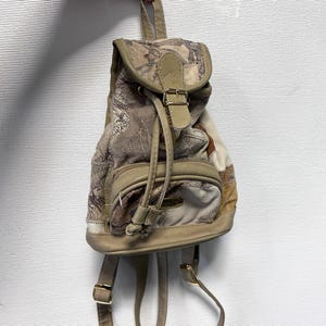 Puede incluir: Una pequeña mochila beige con cierre de solapa y cordón. La mochila presenta un diseño de patchwork con varios patrones y colores, incluyendo beige, marrón y canela. Tiene un pequeño bolsillo frontal y correas ajustables.