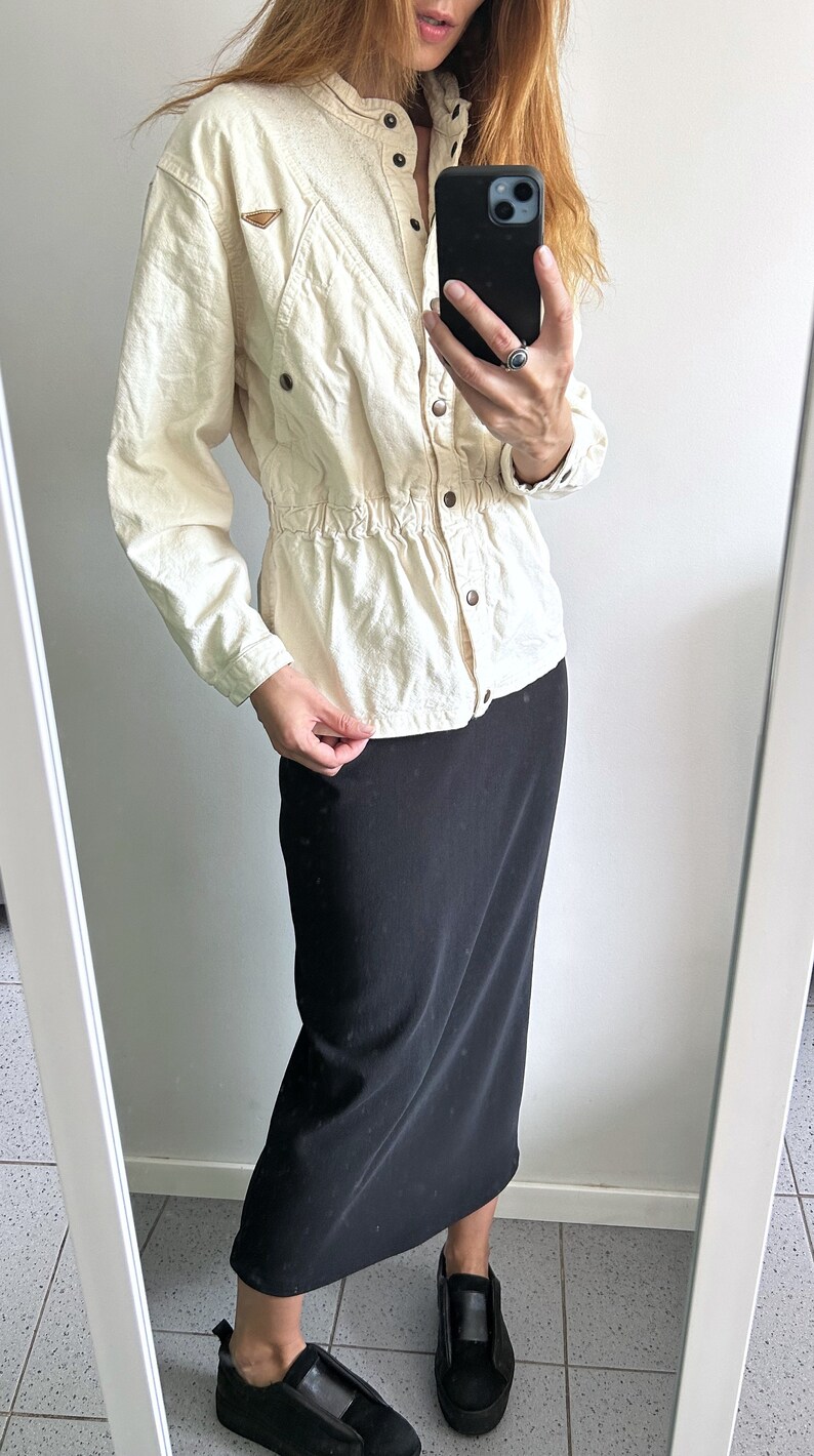 K&ouml;nnte beinhalten: Ein cremefarbenes, lang&auml;rmliges Hemd mit Kn&ouml;pfen, einer eingezogenen Taille und einem braunen Patch auf der linken Seite. Das Hemd wird mit einem schwarzen Midirock und schwarzen Plateauschuhen getragen.