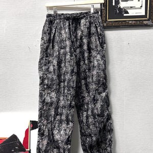 Op de afbeelding: Zwart-witte broek met patroon hangend aan een witte hanger. De broek heeft een taille met trekkoord en een losse pasvorm. De stof heeft een textuur. De broek wordt gepresenteerd tegen een lichtgrijze muur.
