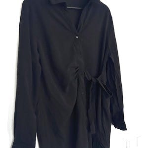 May include: A black long-sleeved shirt dress with a collared neckline and a button-down front. The dress has a tie at the waist and is hanging on a black hanger against a white wall.