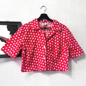 Puede incluir: Una camisa roja de manga corta con un estampado floral blanco. La camisa está colgada en una percha negra contra una pared blanca. La camisa tiene cuello y unos botones en la parte delantera.