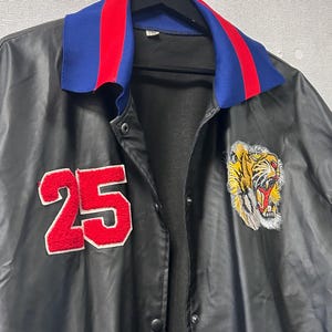 May include: A black jacket featuring a blue and red striped collar, a red felt number 25 patch on the right chest, and an embroidered tiger head patch on the left chest. The jacket has a front snap button closure and a smooth, dark finish.