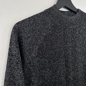 May include: A black long-sleeved top with a high neckline, hanging on a black hanger. The fabric has a textured appearance with silver flecks throughout, creating a shimmering effect. The top is made of a soft, comfortable material.