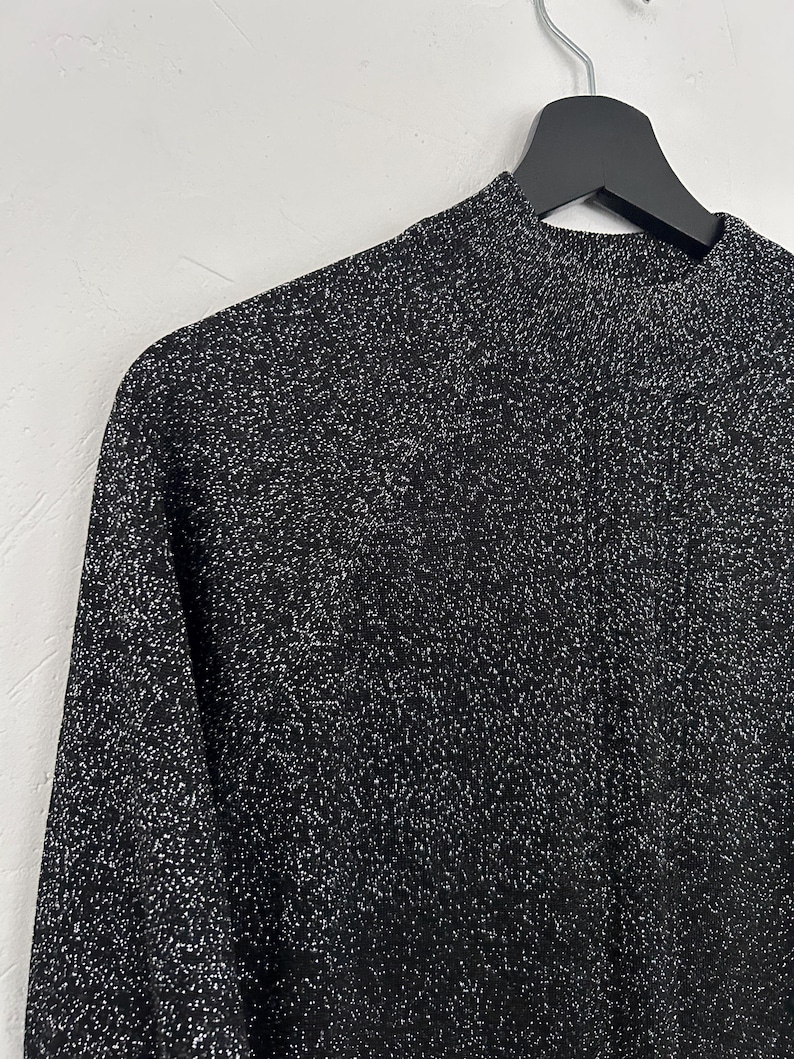 May include: A black long-sleeved top with a high neckline, hanging on a black hanger. The fabric has a textured appearance with silver flecks throughout, creating a shimmering effect. The top is made of a soft, comfortable material.