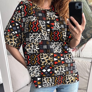 Può includere: Una camicetta a maniche corte con base nera e un motivo patchwork multicolore. Il design include quadrati con stampa leopardata, forme geometriche rosse e bianche, motivi a righe bianche e nere e una scollatura rotonda.