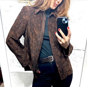 Peut inclure: Une veste marron avec un motif peau de serpent marron foncé, une fermeture éclair frontale intégrale, un col pointu et des manches longues. La veste est portée sur un haut noir à col montant et un pantalon noir avec une ceinture en cuir marron.