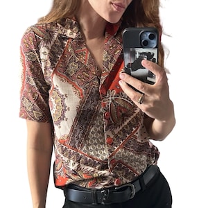 Può includere: Una camicia a maniche corte con bottoni caratterizzata da un vivace motivo paisley e geometrico multicolore nei toni dell'arancione bruciato, crema, marrone e nero. La camicia presenta un colletto classico e un'abbottonatura frontale con bottoni arancioni.
