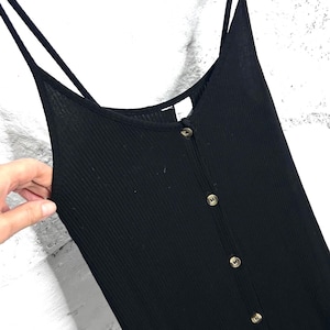Könnte beinhalten: Schwarzes Rippstrickkleid mit dünnen Spaghettiträgern und einer Knopfleiste vorne. Das Kleid hat einen V-Ausschnitt und ist aus einem weichen, dehnbaren Material gefertigt. Die Knöpfe sind hellbraun. Das Kleid hängt an einer Metallstange.