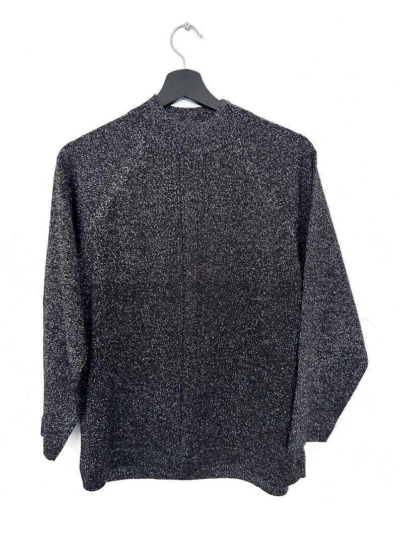 May include: A black and silver speckled jumper with long sleeves and a mock neck, suspended on a black hanger against a white backdrop. The fabric of the jumper has a textured appearance.