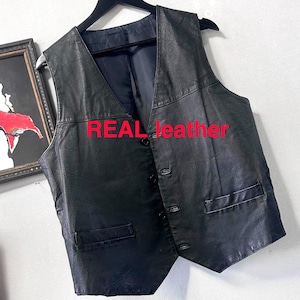 Peut inclure: Gilet en cuir noir avec un col en V et une fermeture boutonnée. Comprend deux poches avant et le texte "REAL leather" en rouge. Le gilet est suspendu à un cintre noir.