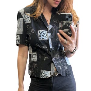 Op de afbeelding: Een zwart overhemd met korte mouwen en knoopsluiting, voorzien van een patchworkontwerp met lichtgrijze en witte dierenprintvierkanten. Het overhemd is versierd met witte geborduurde bloem- en slingerende rankmotieven.