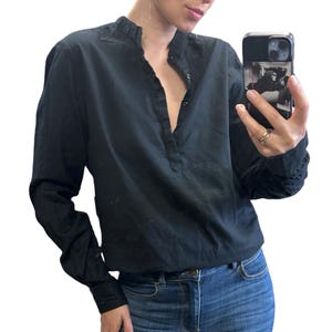 Könnte beinhalten: Ein schwarzes Langarmhemd mit Stehkragen und gerüschtem Rand, einer teilweisen Knopfleiste sowie langen Ärmeln mit Manschetten. Das Hemd wird mit einer blauen Jeans kombiniert, was eine einfache und funktionale Outfit-Kombination ergibt.