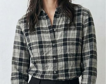 May include: A long-sleeved button-down shirt featuring a black and grey plaid pattern. The shirt has a classic collar, a front button placket with dark buttons, and is paired with solid black trousers. The fabric has a visible woven texture.
