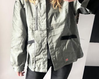 May include: A sage green jacket featuring a vertical zippered chest pocket, two lower front pockets with black trim, a fold-down collar, and a front button closure. The sleeves have black cuffs, and a small rectangular red label is visible on the lower left.