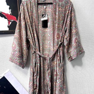 May include: A long-sleeved robe featuring a complex, repeating mandala-inspired pattern in shades of dusty pink, teal, and cream. The garment includes a matching fabric tie belt at the waist, an open front design, and a label reading ANISA.