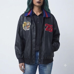 May include: A black bomber jacket featuring a blue collar with red stripes, a tiger head patch on the right chest, and the red number 25 patch on the left chest. The jacket has a snap-button front closure, long sleeves with ribbed cuffs, and a ribbed hem.