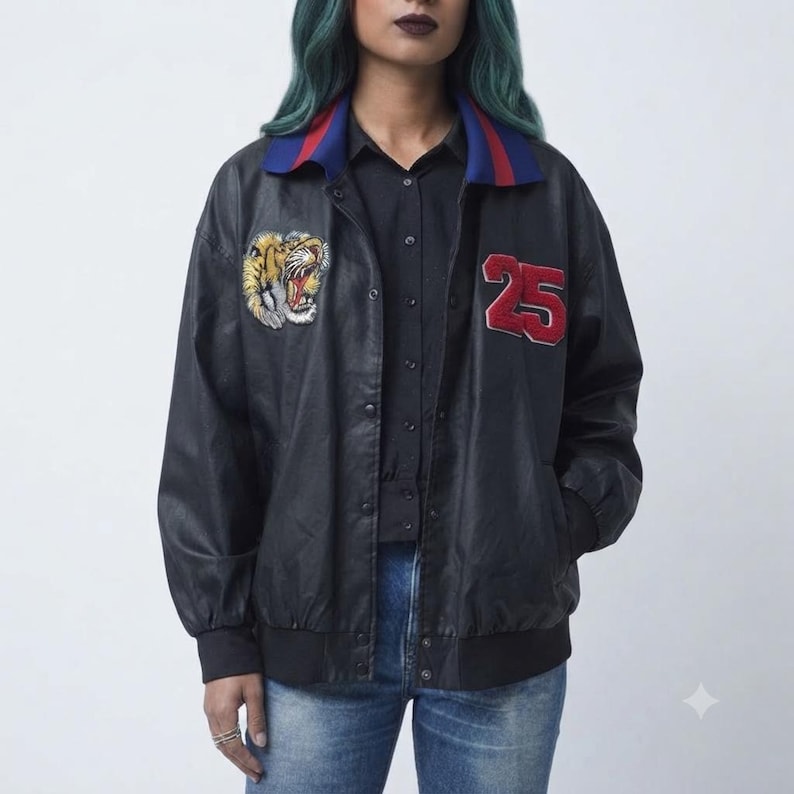 May include: A black bomber jacket featuring a blue collar with red stripes, a tiger head patch on the right chest, and the red number 25 patch on the left chest. The jacket has a snap-button front closure, long sleeves with ribbed cuffs, and a ribbed hem.