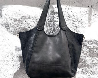 May include: A dark grey leather tote bag with a snakeskin textured side panel. The bag has two long shoulder straps and is hanging against a white textured wall. The bag is a medium size.