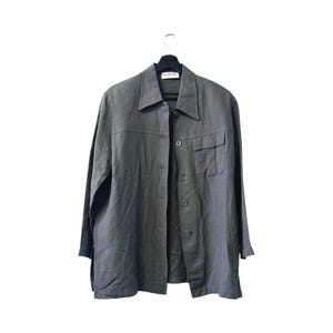 Può includere: Una camicia abbottonata verde oliva scuro con colletto e due tasche sul petto. La camicia è appesa a una gruccia nera su sfondo bianco. Il nome del marchio "monari" è visibile sulla gruccia.