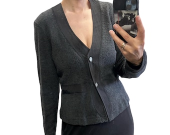 May include: A dark gray long-sleeve cardigan with a textured knit fabric, a deep V-neckline, and a smooth black trim along the front opening. The garment features two round, silver-toned buttons and a horizontal black band across the waist area.